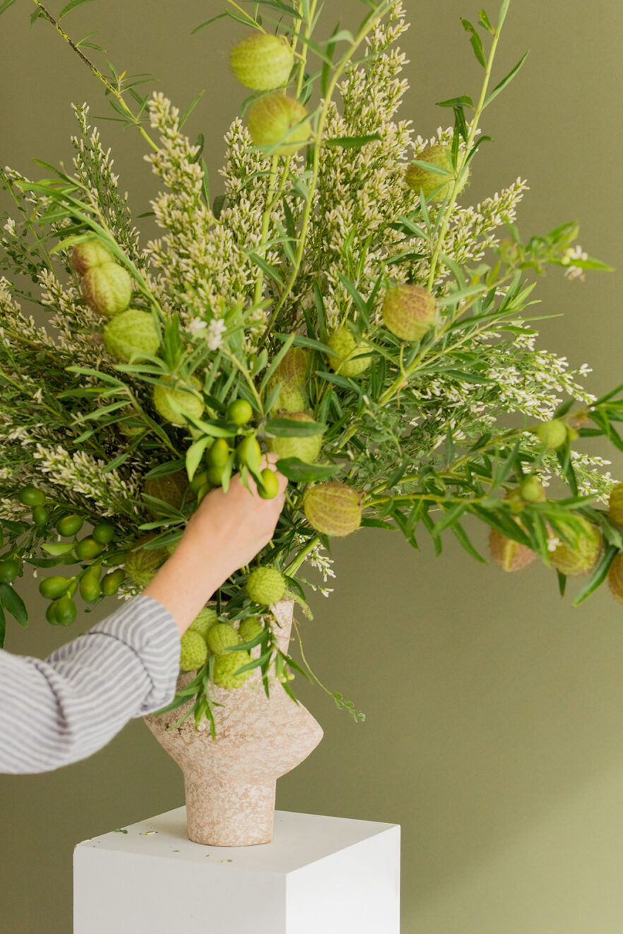 A person arranges green foliage and seed pods in a textured beige vase shaped like a head, set against a plain olive green background.